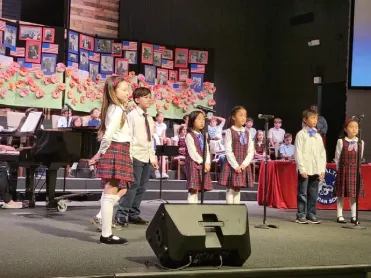 Students in uniforms sing on stage before art displays at a school event or performance.