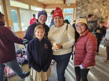 Four people pose and smile indoors; others stand in the background of a lodge-like room.