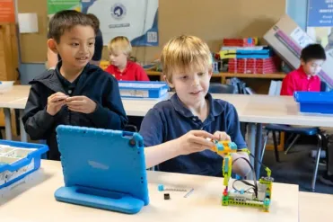 Two boys build a small robot together, smiling, with a blue tablet on their desk.