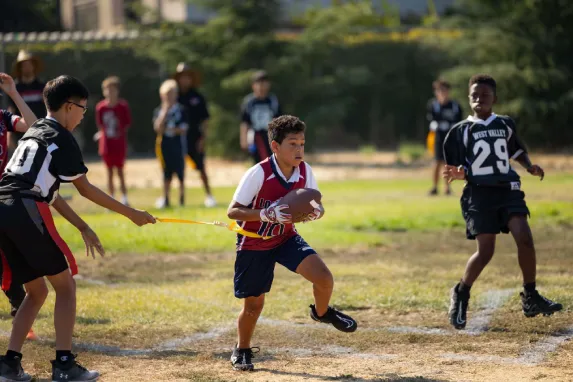 Boy runs with football as another kid grabs his flag; other kids watch in background.