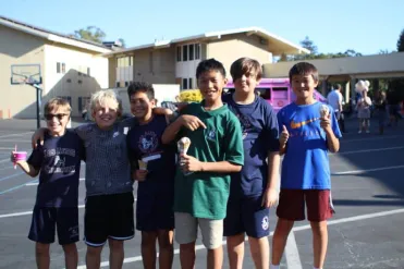 Six boys smiling and holding ice cream stand together outside on a sunny day.